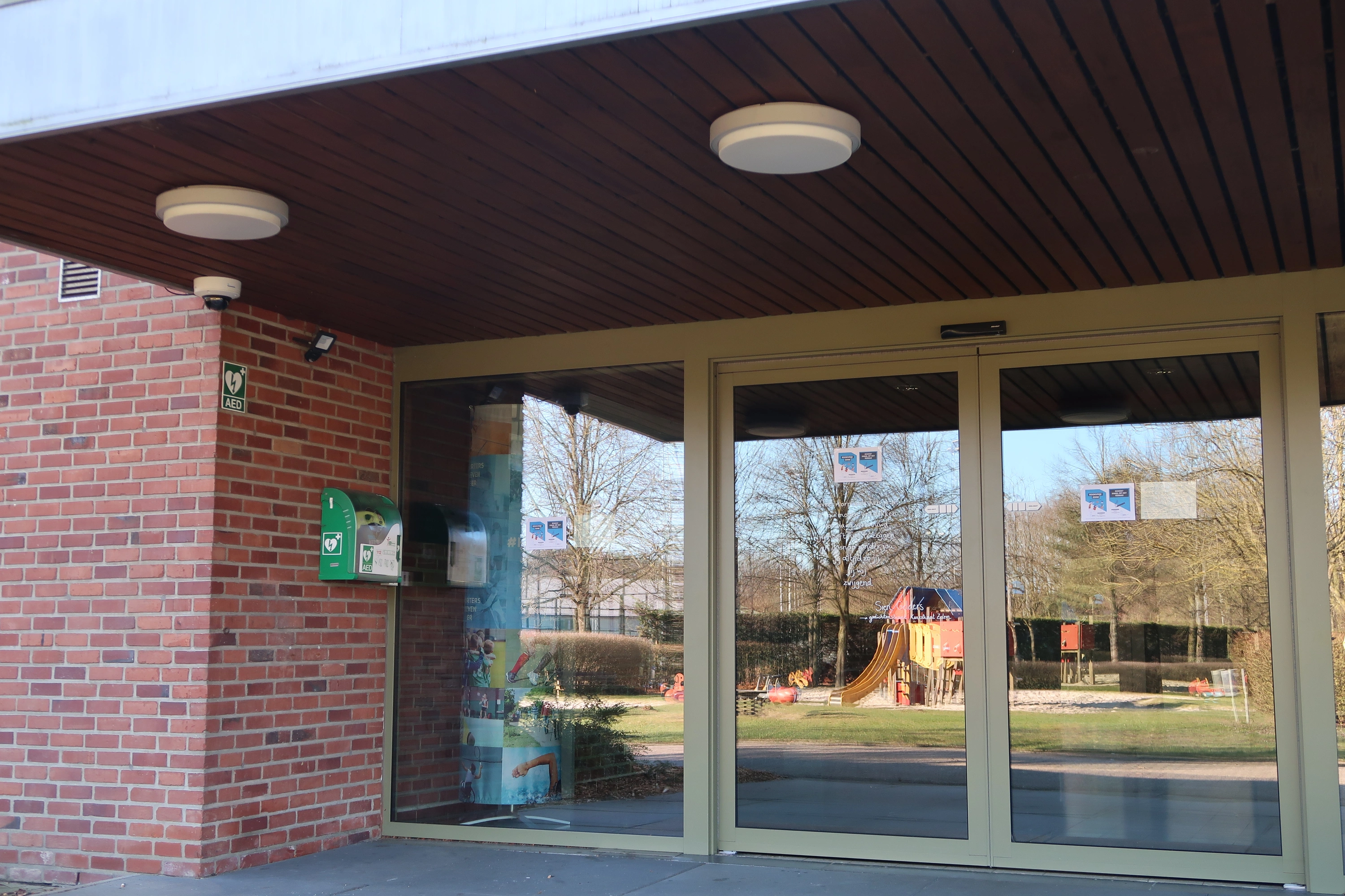 AED Buiten links van de ingangsdeur aan de zijkant van de sporthal, kant van de speeltuin